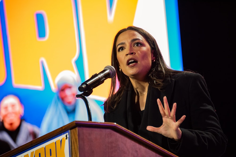 Alexandria Ocasio-Cortez holds her hand out while speaking at a podium, in front of a yellow and blue background.