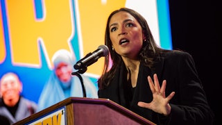Alexandria Ocasio-Cortez holds her hand out while speaking at a podium, in front of a yellow and blue background.