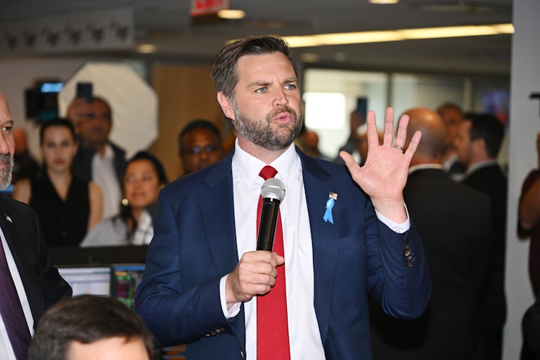 J.D. Vance speaks before a crowd. He has a mic in one hand and raises the other for emphasis.