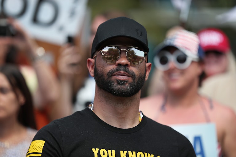 A man wearing sunglasses and a black t-shirt indicating he is carrying a gun stares a head.
