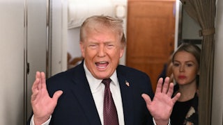 Trump holds up his hands while speaking next to a drawn-aside airplane curtain.