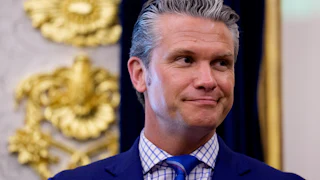 Pete Hegseth standing in the Oval Office wearing a navy suit, blue and white shirt, and blue tie.