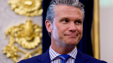 Pete Hegseth standing in the Oval Office wearing a navy suit, blue and white shirt, and blue tie.