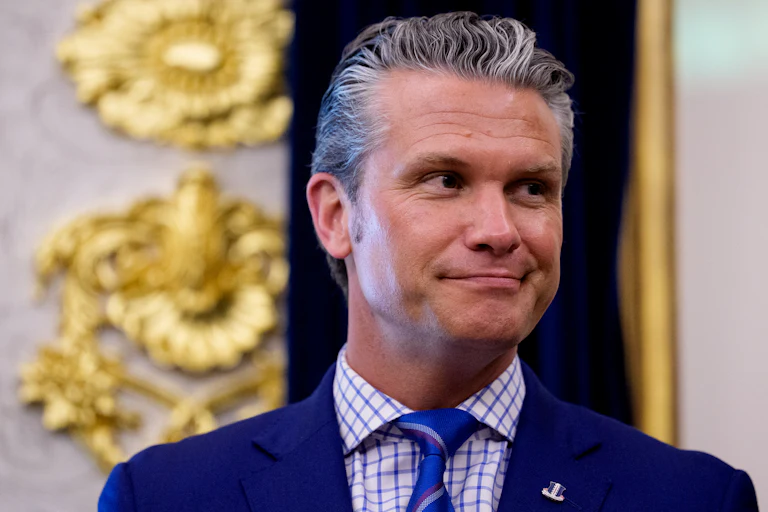 Pete Hegseth standing in the Oval Office wearing a navy suit, blue and white shirt, and blue tie.