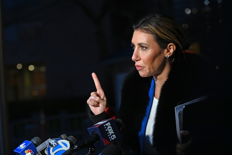 Trump lawyer Alina Habba stands outside at nighttime before several press mics. She points her right index finger to emphasize what she is saying. She appears to be wearing a black fur coat.