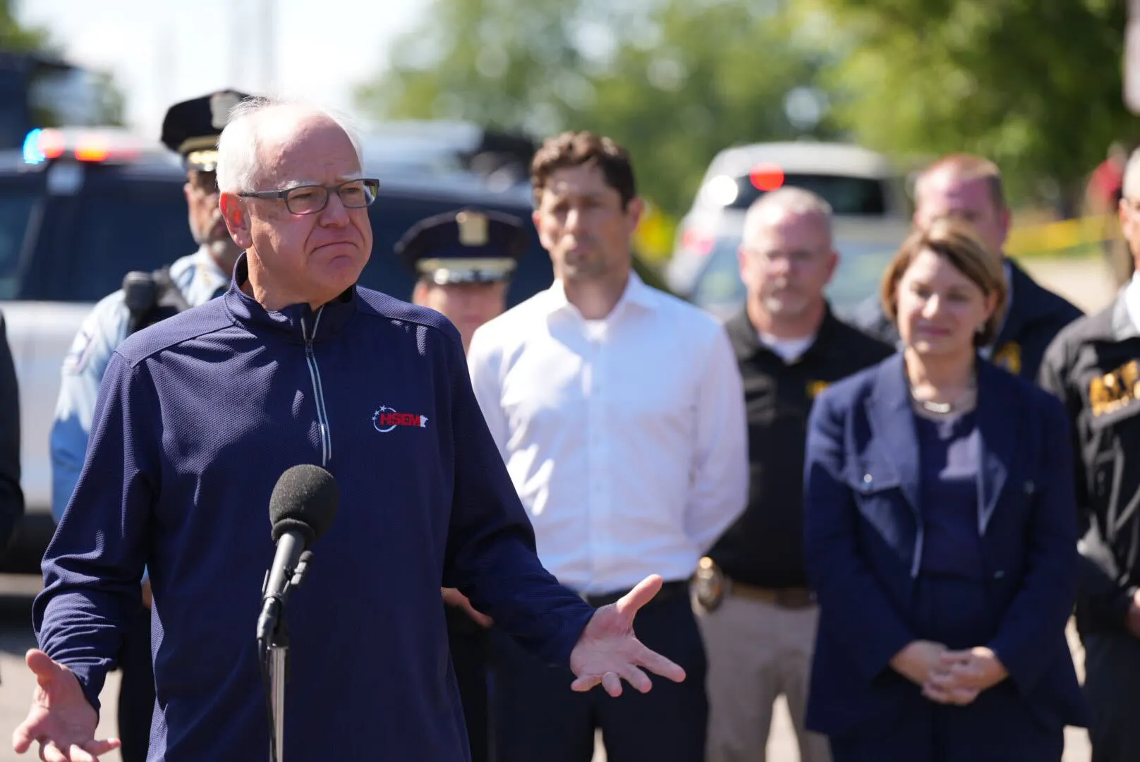 Tim Walz standing outdoors behind a microphone, wearing a windbreaker in front of Minnesota officials including Minneapolis Mayor Jacob Frey and Senator Amy Klobuchar.
