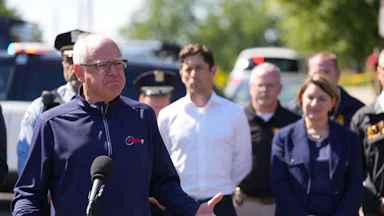 Tim Walz standing outdoors behind a microphone, wearing a windbreaker in front of Minnesota officials including Minneapolis Mayor Jacob Frey and Senator Amy Klobuchar.