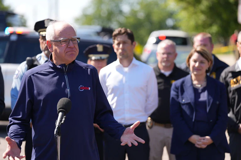 Tim Walz standing outdoors behind a microphone, wearing a windbreaker in front of Minnesota officials including Minneapolis Mayor Jacob Frey and Senator Amy Klobuchar.