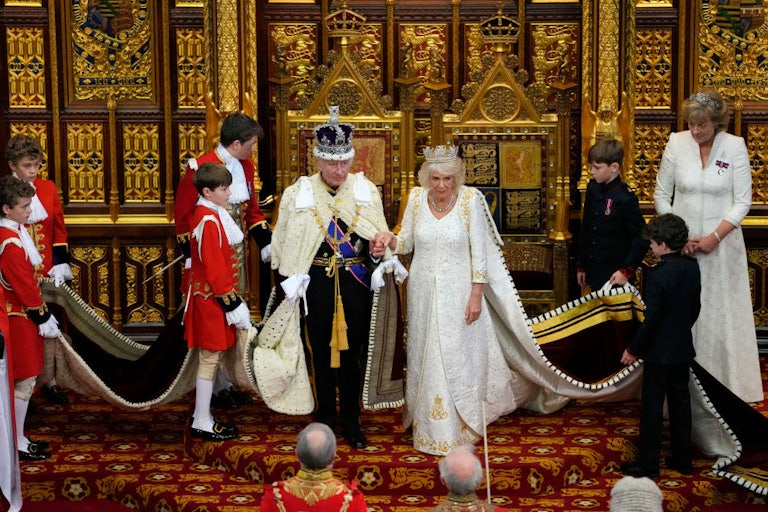 King Charles and Queen Camilla stand in ceremonial attire, surrounded by attendants holding their robes.
