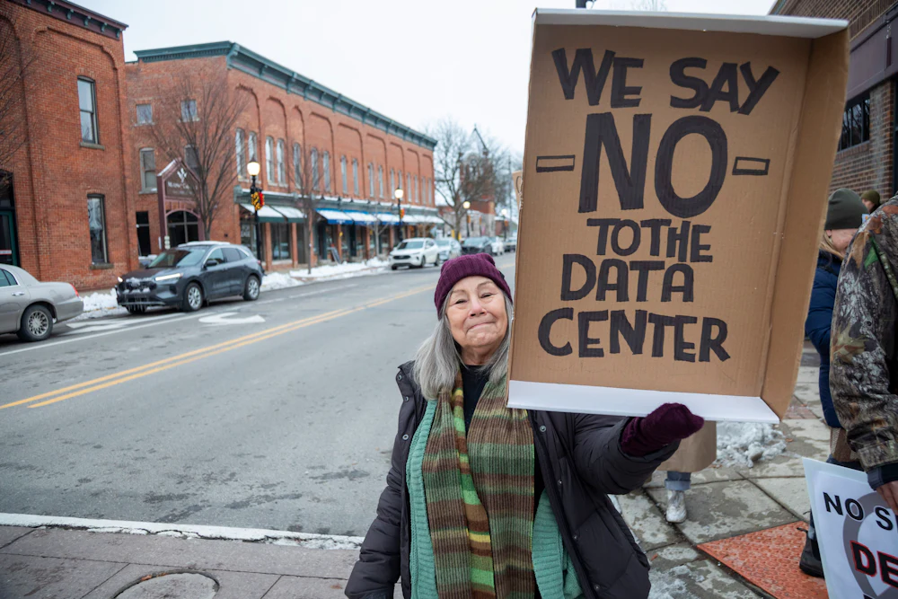 A protester standing with a mostly empty town street in the background holds up a sign saying "We Say NO to the Data Center."