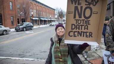 A protester standing with a mostly empty town street in the background holds up a sign saying "We Say NO to the Data Center."