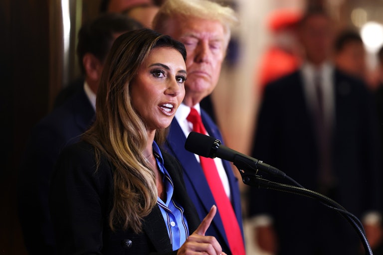 Alina Habba speaks at a microphone. Donald Trump stands to her side and looks on.