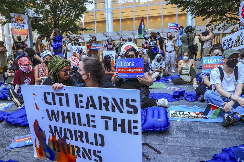 Activists sit with signs with messages such as "Citi Earns While the World Burns," "Citibank: A Partner in Oppression," "Stop Funding Fossil Fuels," and "Stop Funding Genocide."