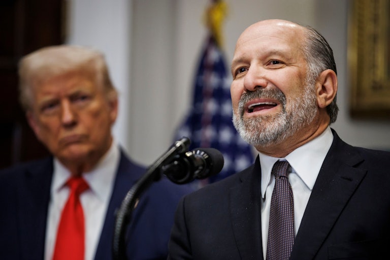 Howard Lutnick speaks into a microphone in the White House while Donald Trump looks on in the background.
