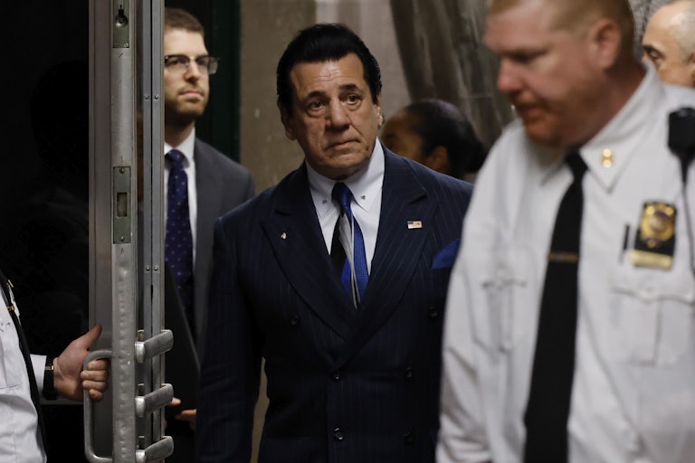 Chuck Zito, wearing a suit and blue tie, enters a room as a door is held open for him. A security guard stands in front of him. Others are in the background.