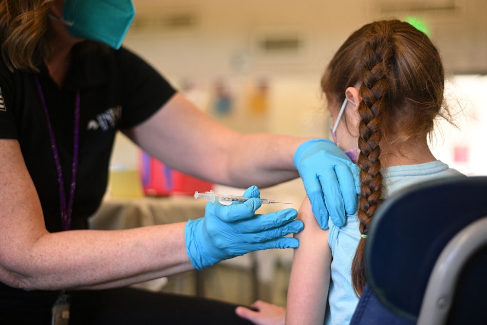 A person wearing blue gloves holds a syringe and needle up to the arm of a child facing away from the camera.