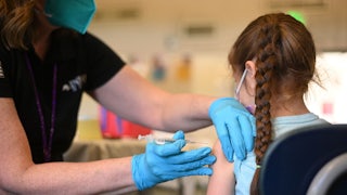 A person wearing blue gloves holds a syringe and needle up to the arm of a child facing away from the camera.