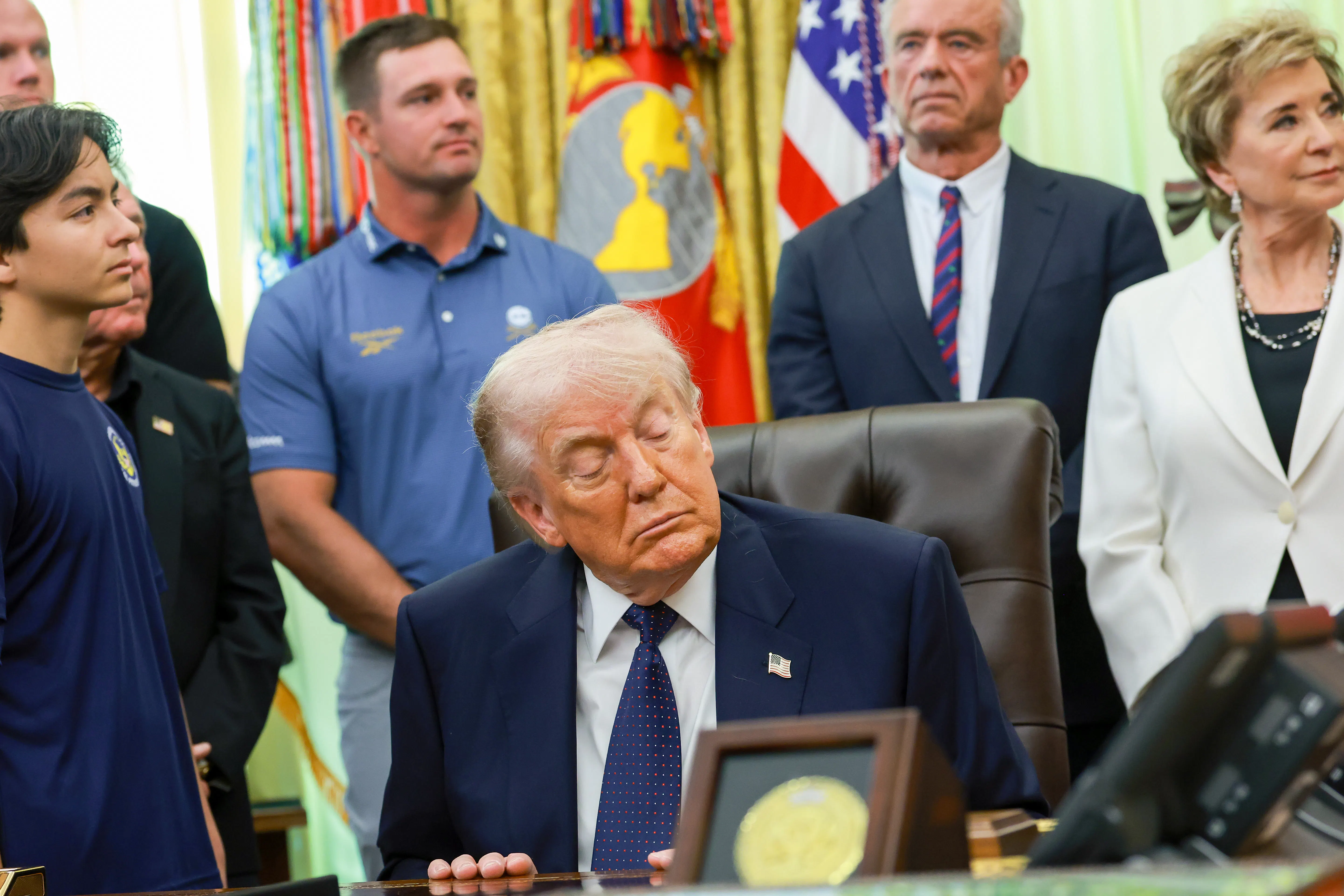 President Donald Trump falls asleep while sitting at his desk in the Oval Office of the White House. Cabinet officals and kids stand around him.