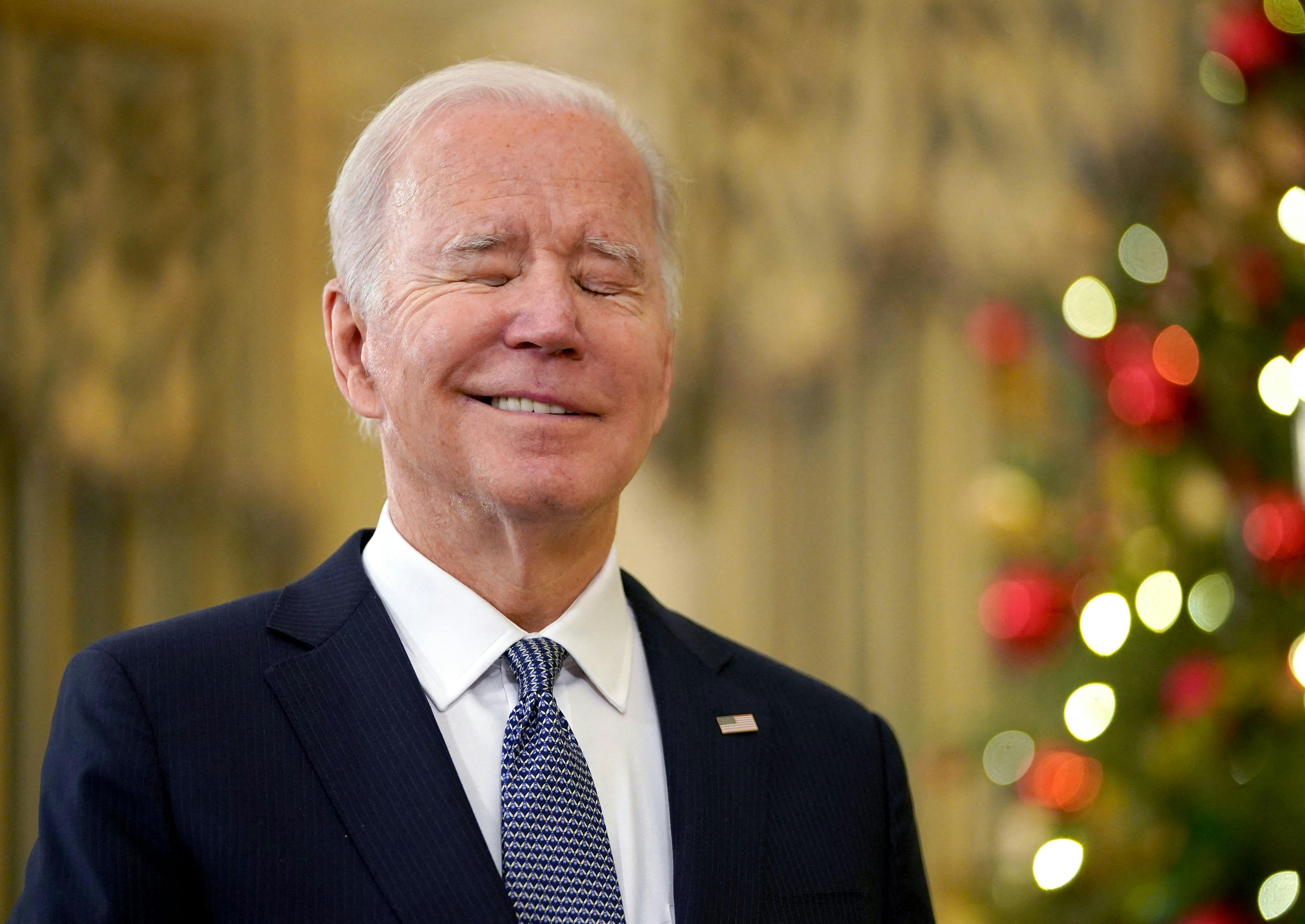 Joe Biden closes his eyes and does a wry little smile as he stands in front of a Christmas tree