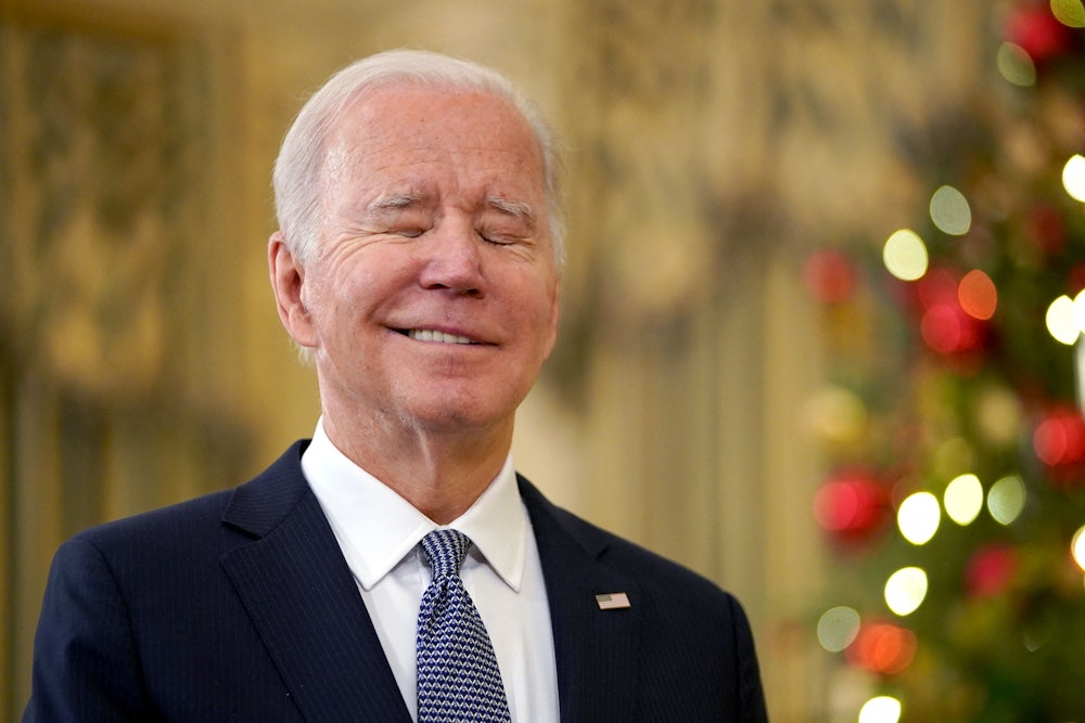 Joe Biden closes his eyes and does a wry little smile as he stands in front of a Christmas tree