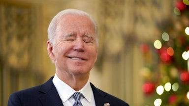Joe Biden closes his eyes and does a wry little smile as he stands in front of a Christmas tree
