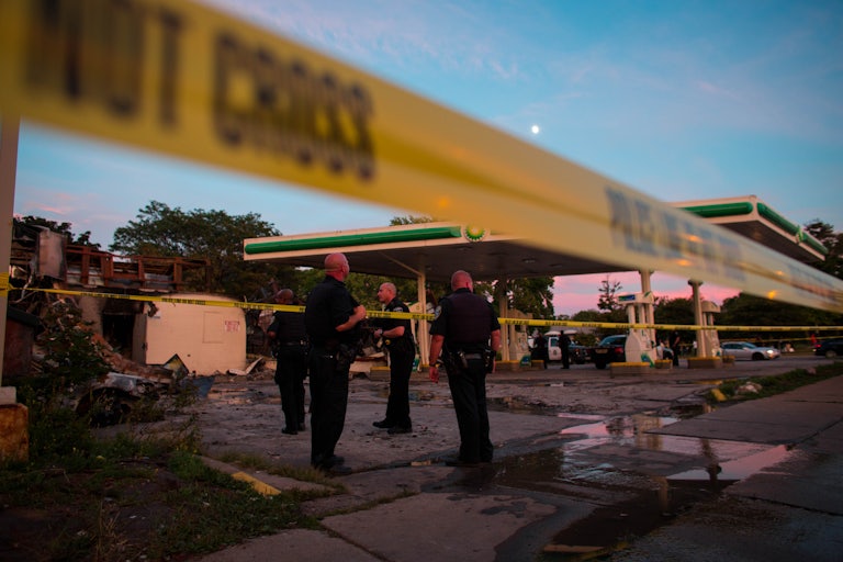 Police officers, gathered behind a police tape line, survey a burned out gas station 