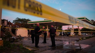 Police officers, gathered behind a police tape line, survey a burned out gas station