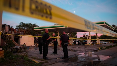 Police officers, gathered behind a police tape line, survey a burned out gas station