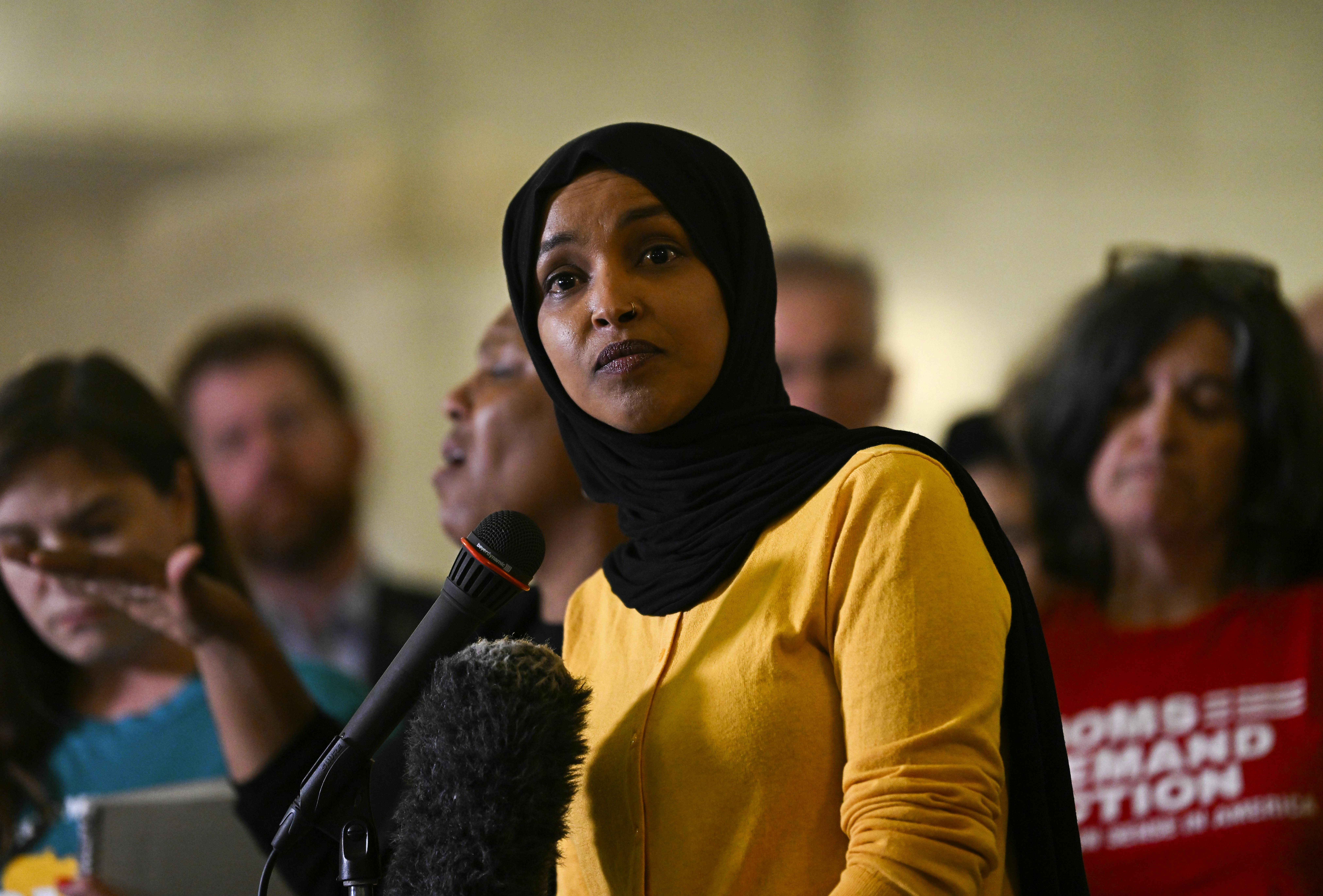 Representative Ilhan Omar speaks at a mic as others stand behind her.