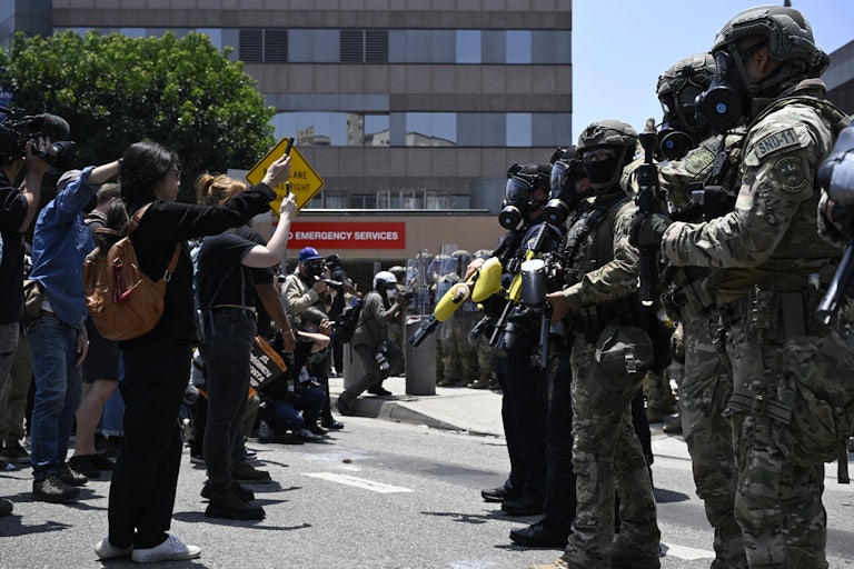 A line of ICE officers, covered head to toe and wearing gace masks, stands in front of a line of protesters, many of them holding phones up pointed at the officers.