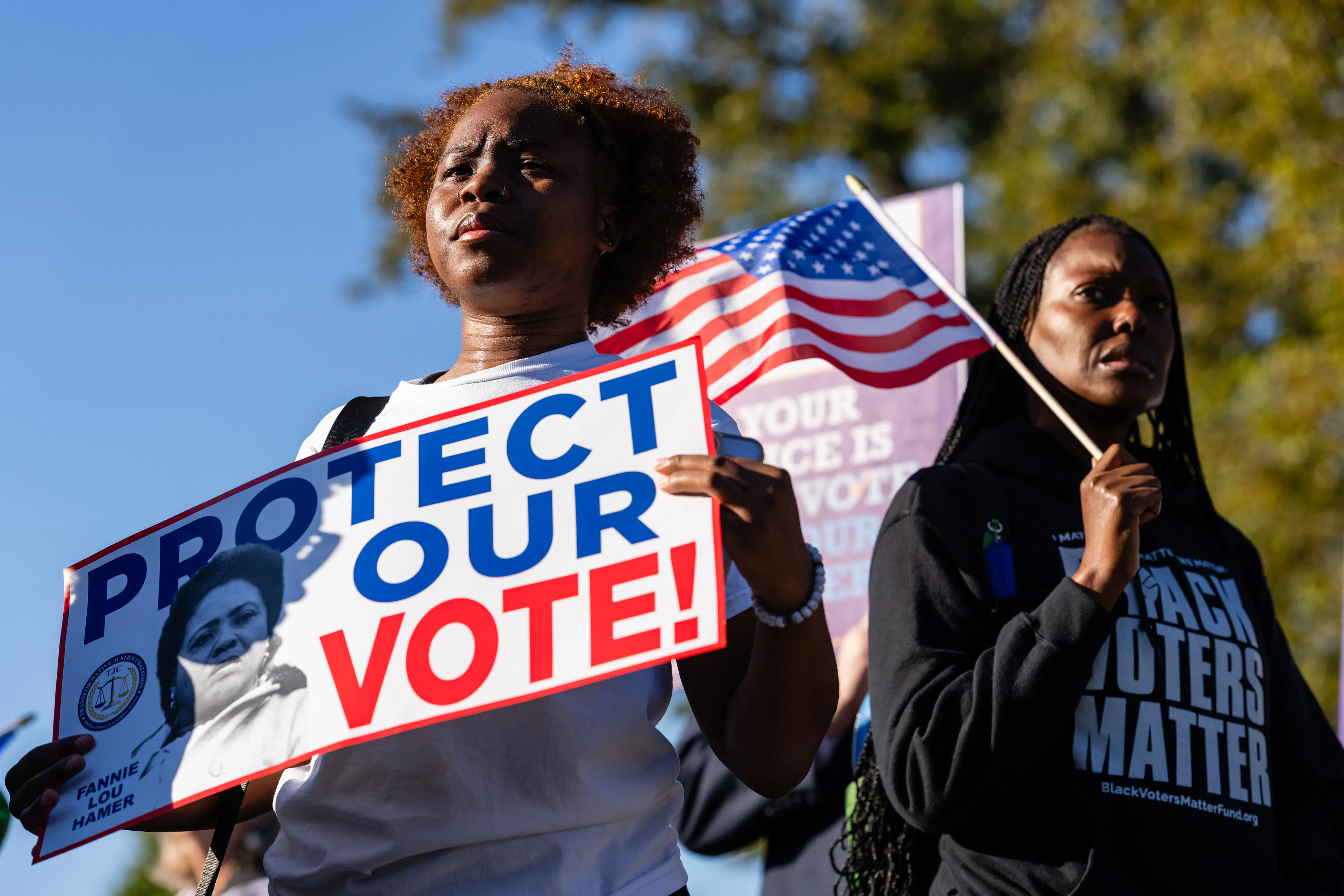 Black demonstrators outside the Supreme Court. One holds up a sign that reads "Protect Our Vote" and another holds an American flag and wears a shirt that says "Black Voters Matter."