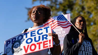 Black demonstrators outside the Supreme Court. One holds up a sign that reads "Protect Our Vote" and another holds an American flag and wears a shirt that says "Black Voters Matter."