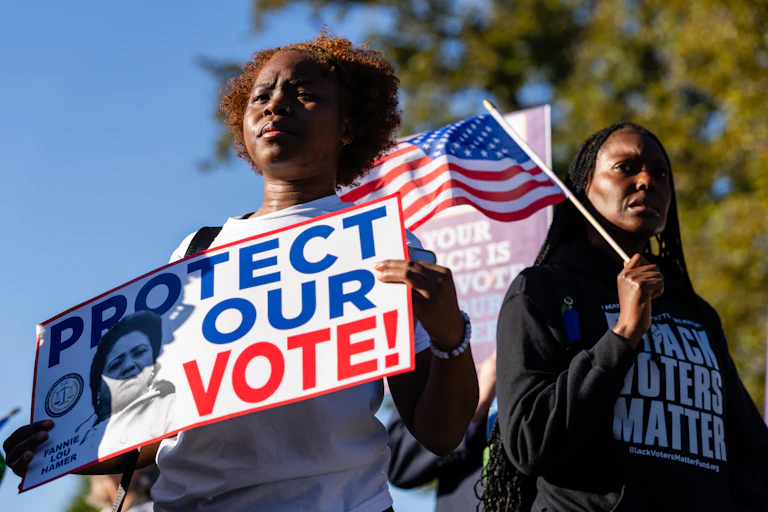 Black demonstrators outside the Supreme Court. One holds up a sign that reads "Protect Our Vote" and another holds an American flag and wears a shirt that says "Black Voters Matter."