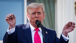 Donald Trump speaks at the presidential podium and raises both hands as if conducting an orchestra.