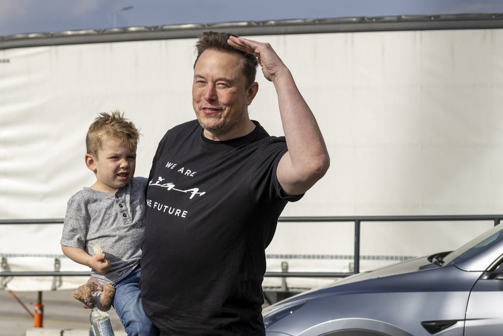 Elon Musk holds a small child while wearing a black t-shirt that reads "We Are The Future."