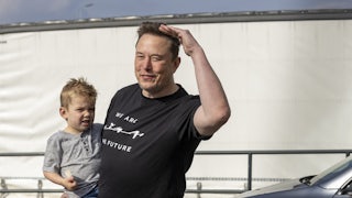 Elon Musk holds a small child while wearing a black t-shirt that reads "We Are The Future."