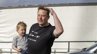 Elon Musk holds a small child while wearing a black t-shirt that reads "We Are The Future."