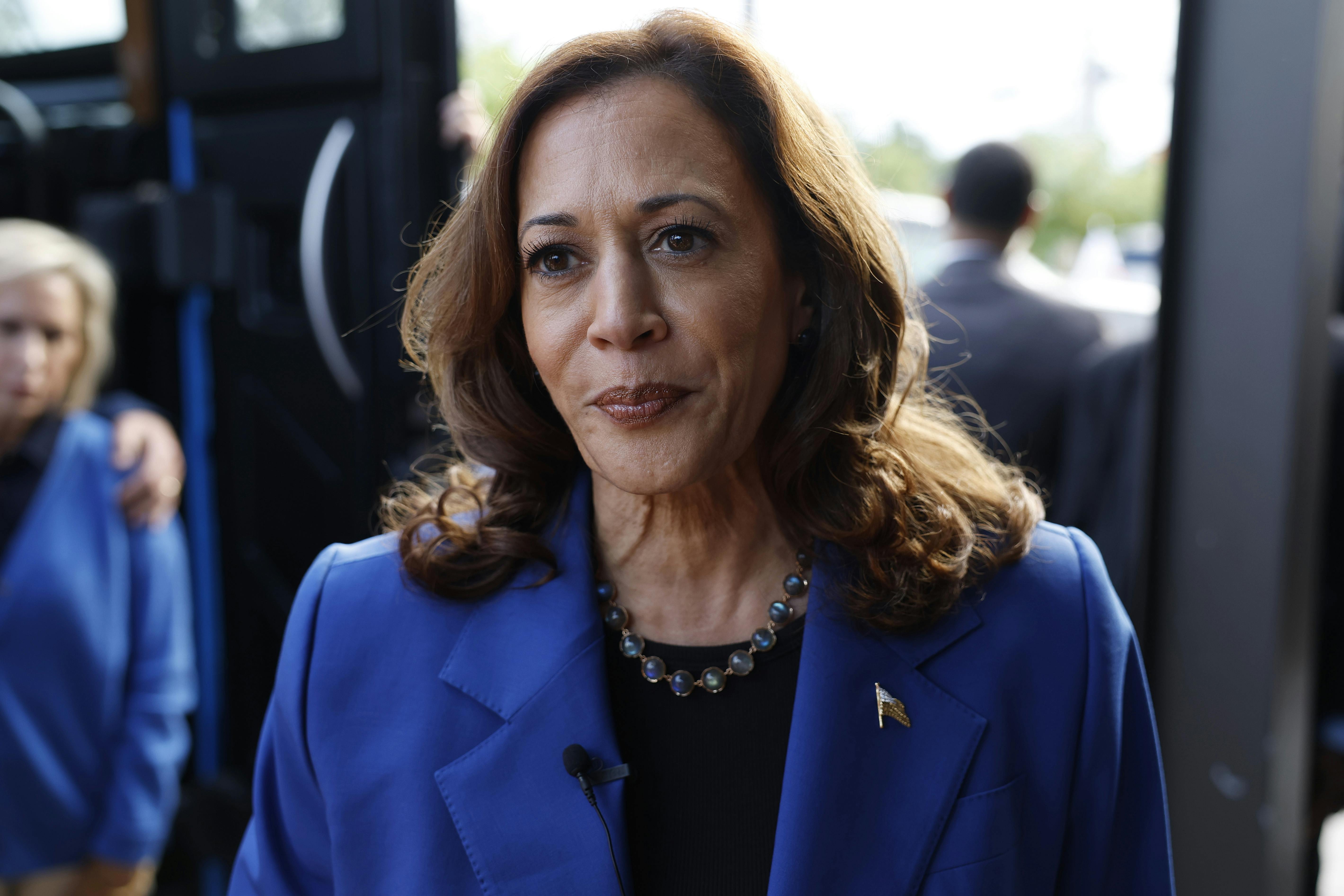 Kamala Harris, wearing a necklace and blue pantsuit, stands outside a bus and purses her lips as she speaks to reporters. 