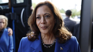 Kamala Harris, wearing a necklace and blue pantsuit, stands outside a bus and purses her lips as she speaks to reporters.