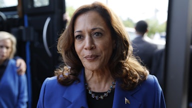 Kamala Harris, wearing a necklace and blue pantsuit, stands outside a bus and purses her lips as she speaks to reporters.