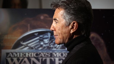 John Walsh, in profile, stands in front of a poster for “America’s Most Wanted”