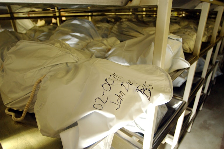 Body bags lie on shelving, the front one labeled "John Doe."