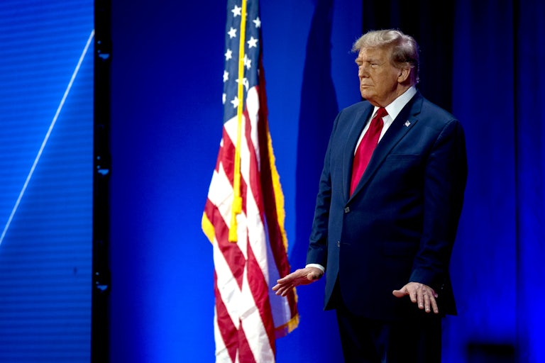 Donald Trump stands with his hands flared out next to a large American flag