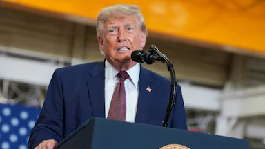 Donald Trump stands at a podium before a microphone with the presidential seal wearing a blue suit and tie with a faint American flag partially visible beind him.