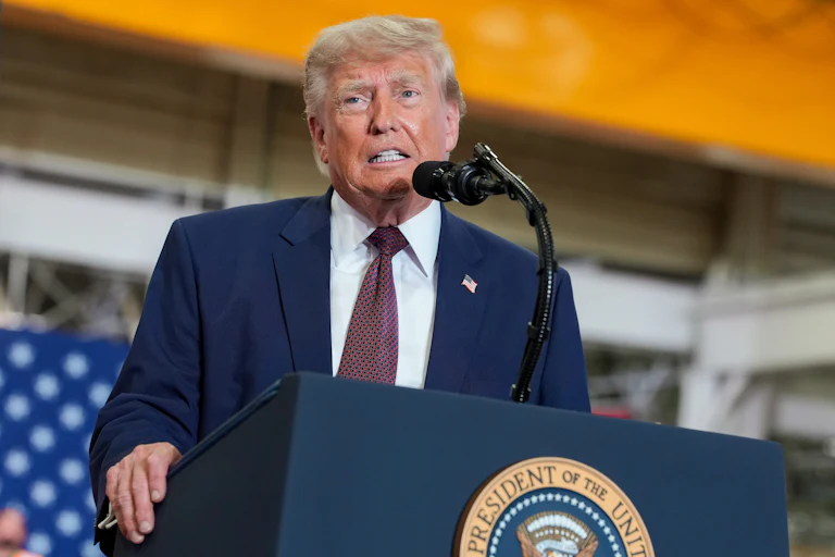 Donald Trump stands at a podium before a microphone with the presidential seal wearing a blue suit and tie with a faint American flag partially visible beind him.
