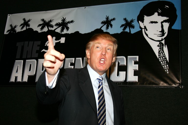 Donald Trump stands in front of an advertisement for "The Apprentice." He is pointing outward and speaking his catchphrase "You're fired."