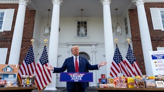 Trump holds his hands out between tables filled with cereal, model houses, mustard, and other products, while standing at a lectern.