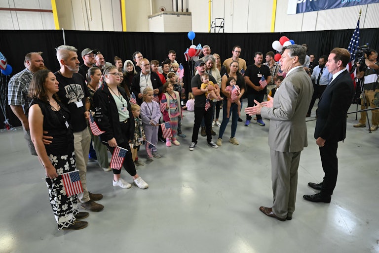 A group of white adults and children hold U.S. flags as they listen to two Trump officials speak.
