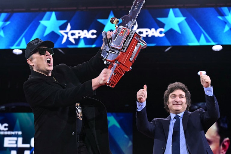 Elon Musk waves a chainsaw above his head on the CPAC stage. Another white man behind him holds two thumbs up in the air.