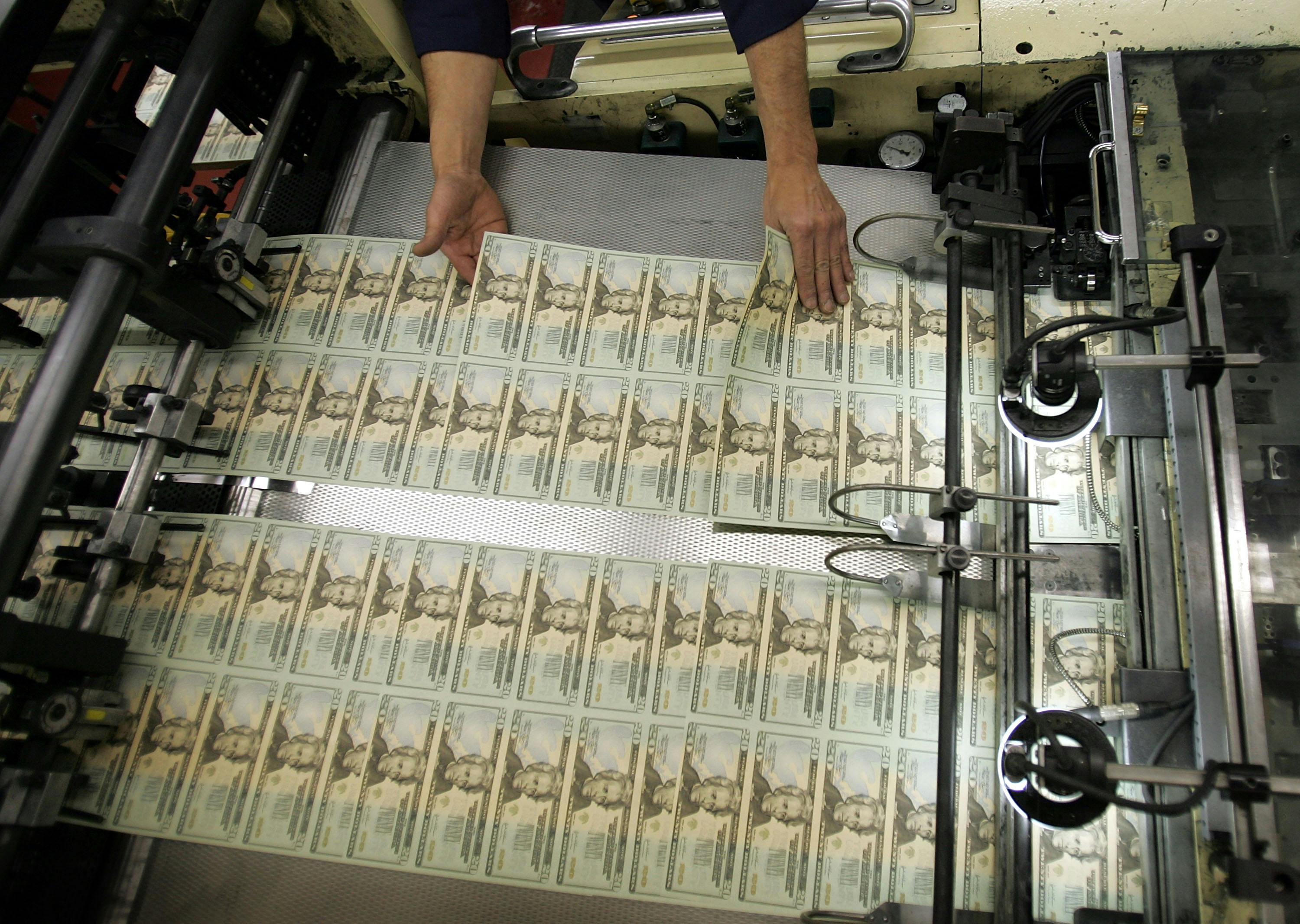 A worker's hands hover over recently printed twenty-dollar bills, inspecting them.